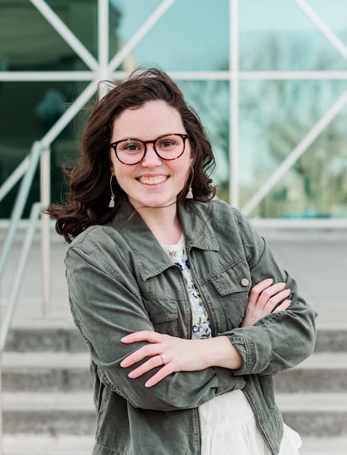Portrait of photographer, Shelby Rae, smiling with arms crossed in front of a modern glass building.