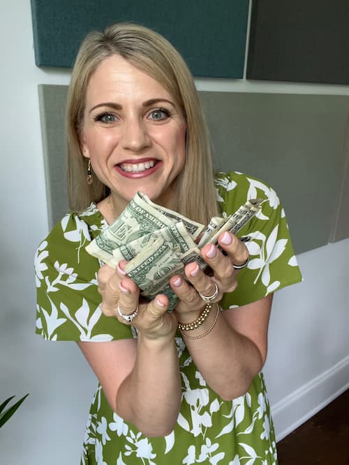 A smiling woman in a green floral dress holding a large handful of crumpled dollar bills toward the camera.