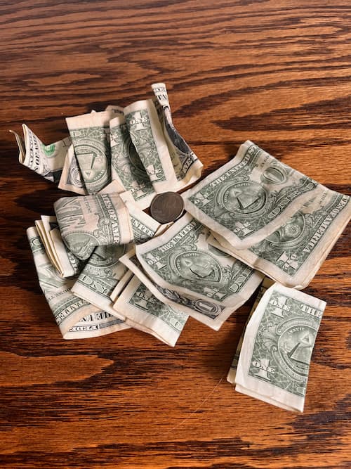 A messy pile of crumpled one-dollar bills and a single coin scattered on a wooden surface. The disorganized cash visually represents financial chaos and the struggle behind finding it difficult to save money.