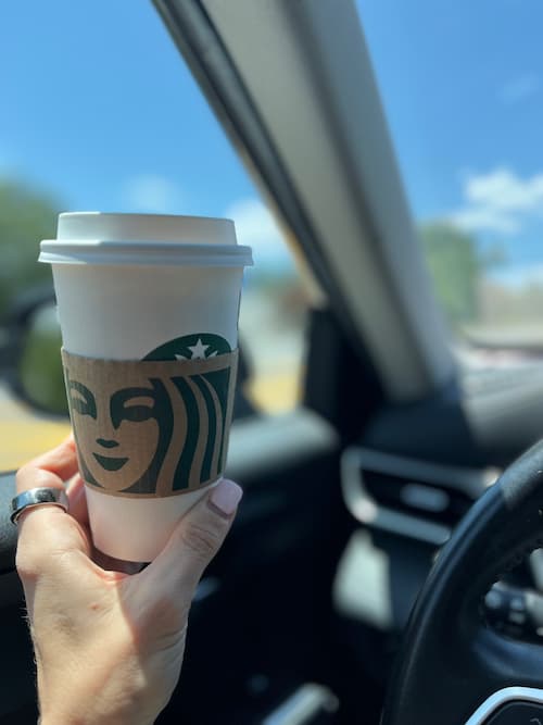 A hand holding a Starbucks coffee cup inside a car, with sunlight streaming through the window and a clear blue sky outside. The image subtly illustrates a common example of everyday splurges, aligning with themes around how to stop impulsive spending.